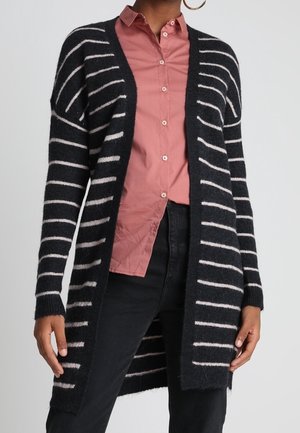 Cardigan long à rayures noires et blanches porté sur une chemise rose à boutons et un pantalon noir, porté par une personne avec des boucles d'oreilles créoles.