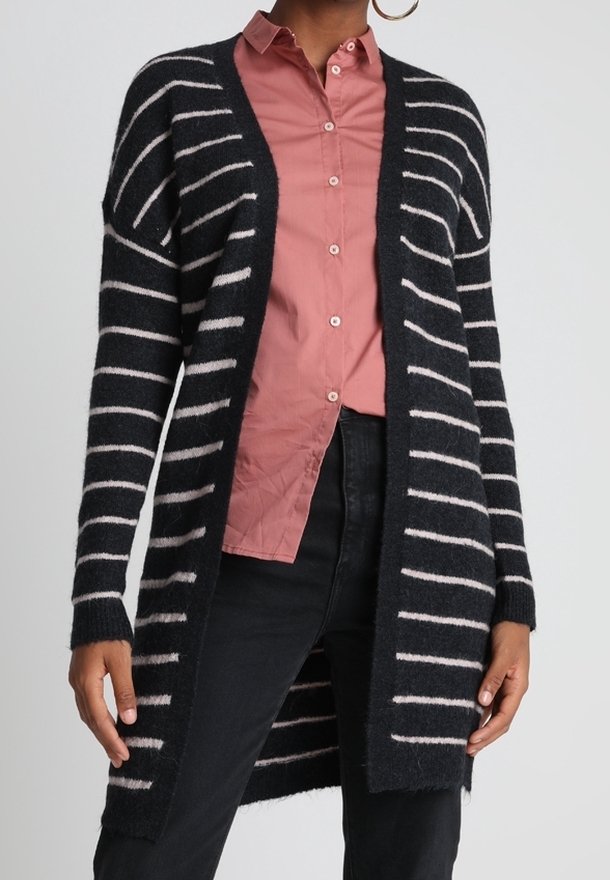 Cardigan long à rayures noires et blanches porté sur une chemise rose à boutons et un pantalon noir, porté par une personne avec des boucles d'oreilles créoles.