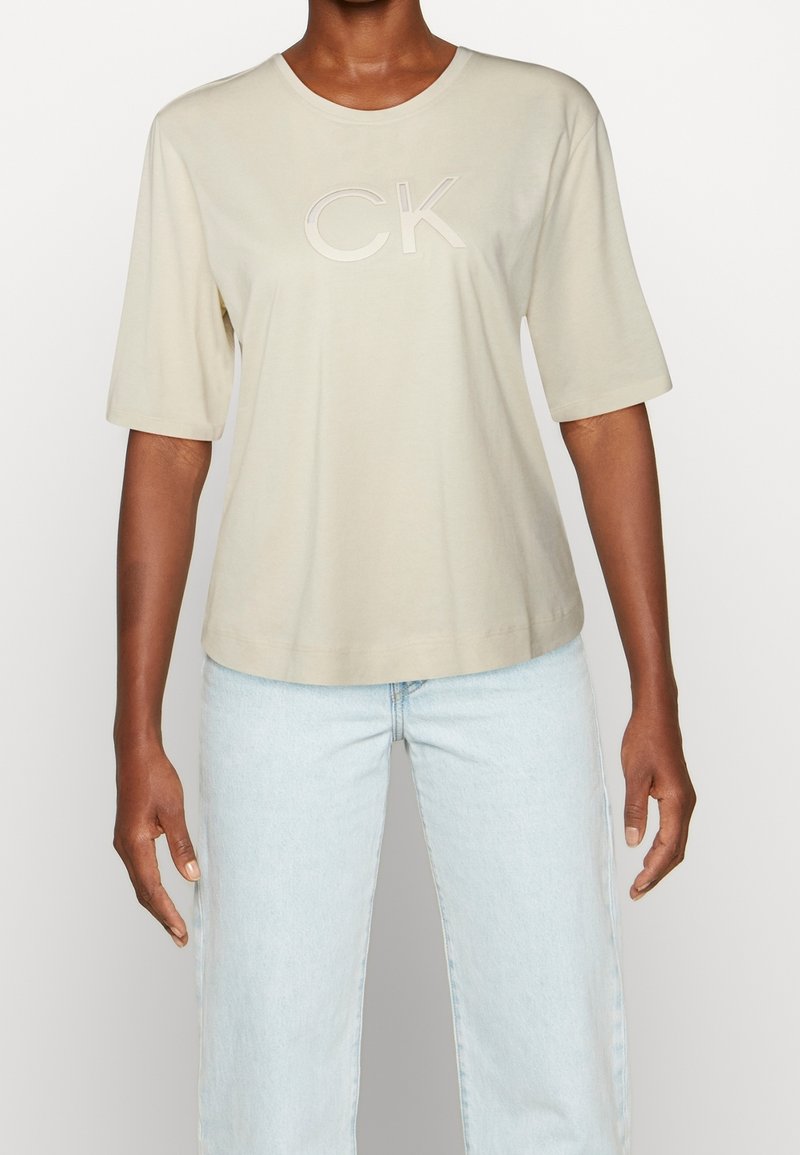 Beige bomulds t-shirt med korte ærmer, rund halsudskæring og præget "CK" logo på brystet. Parret med lyseblå jeans.
