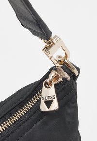 Sort nylon taske med guldfarvede hardware, prydet med en lynlås med "GUESS" indgravering og en klipslukning. Glat tekstur, kompakt design.