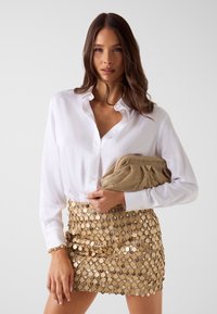 Witte blouse met V-hals, gecombineerd met een gouden pailletten mini rok met cirkelvormige pailletten. Het model houdt een gestructureerde gouden clutch vast.