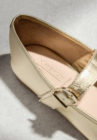 Béžová balerína se zlatou přezkou a měkkou vnitřní výstelkou, zkosená na světle šedém texturovaném povrchu.