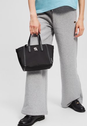 Sac à main en nylon noir avec deux poignées courtes, présentant des détails en métal argenté et des coutures contrastées, posé à côté d'un pantalon de survêtement gris.