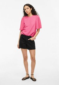 Top rosa texturizzato con maniche corte, abbinato a pantaloni corti neri. La modella indossa ballerine in rete nere, in piedi contro uno sfondo bianco uniforme.