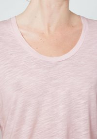 Gaiši rozā īsām piedurknēm T-krekls ar teksturētu apdari un plašu izgriezumu. Audums izskatās mīksts un nedaudz caurspīdīgs.