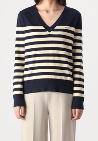 Granatowo-kremowy sweter w paski z dekoltem w serek, długimi rękawami, ściągaczami przy mankietach i małym logo na piersi. Miękka dzianina.