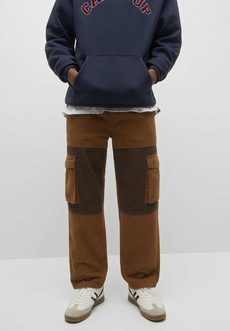 Person iført brune cargo-bukser med mørkere knælapper, en navy blå hættetrøje og hvide sneakers med sorte striber. Hænderne i lommerne.