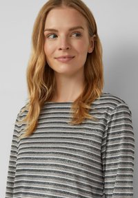 Vrouw met lang blond haar, gekleed in een grijs en zwart horizontaal gestreept topje, kijkt iets naar links tegen een effen lichte achtergrond.