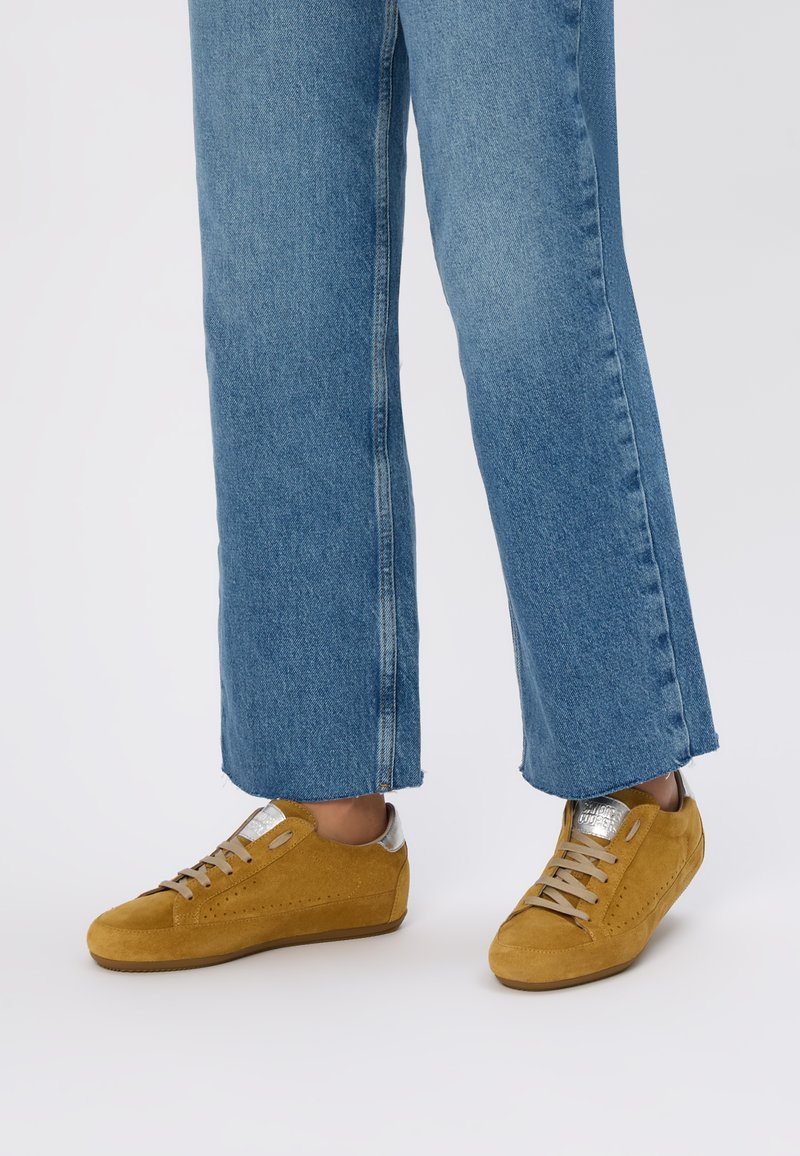 Sneaker in suede giallo con accenti argentati, punta arrotondata e design perforato. Indossati con pantaloni in denim blu a vita corta, con orlo sfrangiato.