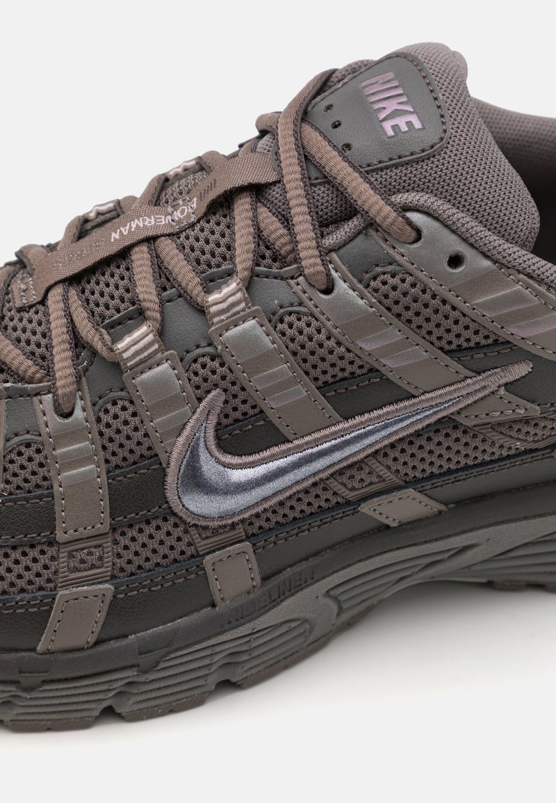 Zapatilla deportiva de color marrón oscuro que cuenta con detalles de malla y cuero, un destacado logo de Nike en plata, y una suela de goma texturizada.