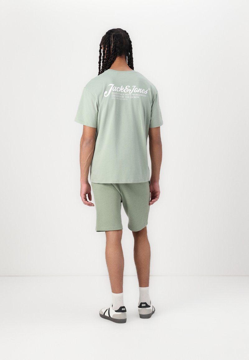 T-shirt a maniche corte, di cotone leggero verde chiaro con logo bianco sulla schiena, abbinato a pantaloni corti coordinati, indossato con sandali a fascia neri e bianchi.