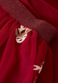 Tessuto rosso con cintura glitterata e faccia di renna sorridente ricamata parzialmente visibile sotto strati morbidi di rete.
