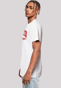 White cotton t-shirt with short sleeves, featuring red printed text on the front. Model stands sideways, showcasing a relaxed fit.