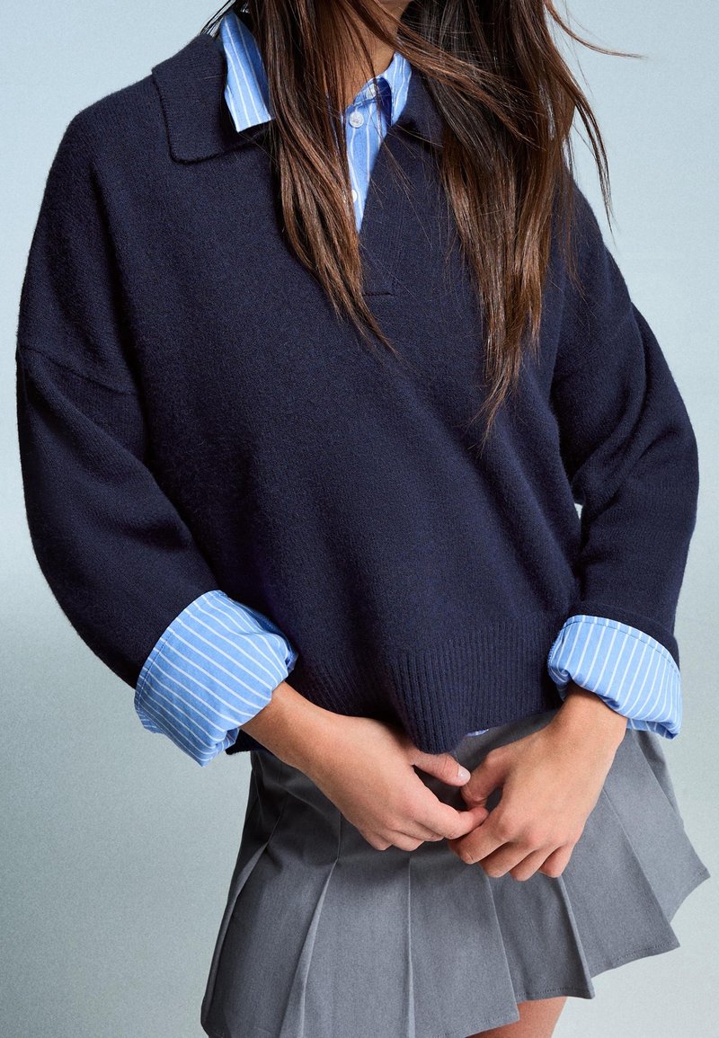 Personne portant un pull bleu marine par-dessus une chemise rayée bleue, associé à une jupe plissée grise, les mains jointes à la taille.