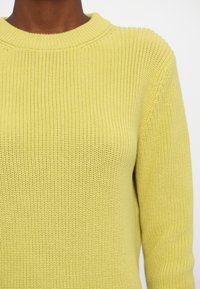 Strikket genser i lys gul med rund hals, med en teksturert overflate og langermede ermer. Glatt stoff med ribbestrikkede detaljer.