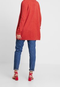 Röd lång kofta med avslappnad passform, strukturerad stickning och ribbade manschetter, i kombination med blå denimjeans och röda klacksandaler med ankelband.