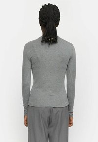 Šedé tričko s dlouhým rukávem, střih těsně přiléhající k tělu, hladká textura a zaoblený lem, nošené s světle šedými kalhotami, pohled zezadu.