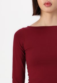 Burgundy long-sleeve top with a boat neckline, made of a smooth fabric, featuring a fitted silhouette and minimal stitching details.