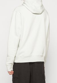 Ljusgrå hu hoodie med mjuk textur, känguruficka och ribbade muddar, bärs med mörka cargobyxor, visar bakifrån.