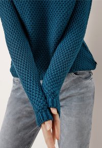 Tealowy sweter z dzianiny o fakturowanym wzorze, z prążkowanymi mankietami i lekko luźnym krojem. Połączony z jasnoszarymi dżinsami, eksponując rękawy i fason.