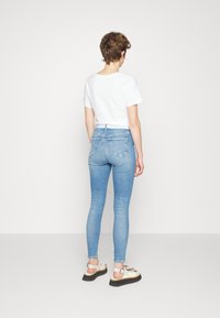 Osoba oblečená v světle modrých skinny džínách, bílém zkráceném tričku a bílých sandálech s černými podrážkami, stojící zády k objektivu na jednobarevném pozadí.