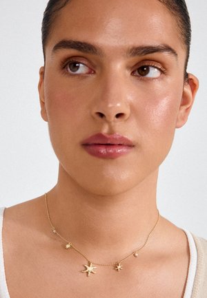 Woman with smooth skin and dark hair, wearing a delicate gold necklace with star and pearl charms, looking slightly to her left.