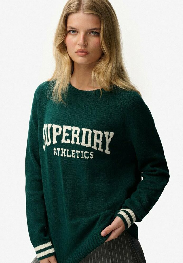 Młoda kobieta z długimi blond włosami, ubrana w ciemnozielony sweter z napisem "SUPERDRY ATHLETICS" i paskowanymi mankietami, stojąca na białym tle.