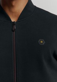 Zwarte ritsjack met een gestructureerde stof, met een bordeauxrode rits en een hexagonaal logo op de linkerborst.