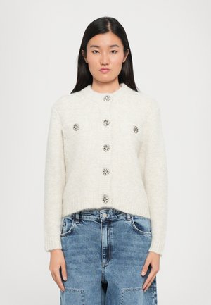 Vaalean kerma cardigan pyöreällä kaula-aukolla, viidellä koristepainikkeella ja kahdella etutaskulla, jossa on ribbejä hihansuissa ja helmassa. Yhdistetty vaaleansinisiin farkkuihin.