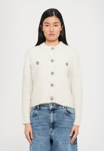 Cardigan crème clair avec un col rond, cinq boutons décoratifs et deux poches avant, avec des poignets et un ourlet côtelés. Porté avec un denim bleu clair.