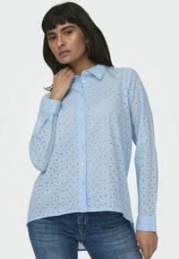 Camicia azzurro chiaro a maniche lunghe con colletto a bottoni, caratterizzata da un sottile motivo a pois e una curvatura dell'orlo, indossata sopra un paio di jeans scuri.