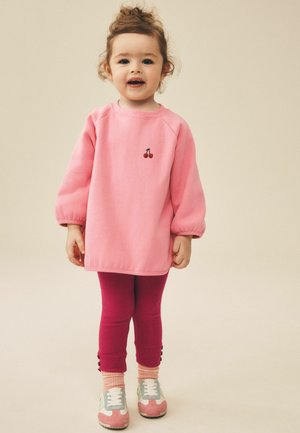 Kleinkind trägt ein rosa Langarmshirt mit Kirschmuster, dunkelrosa Leggings mit Knöpfen und rosa Socken mit Turnschuhen, steht auf einem beigen Boden.