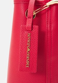 Czerwona skórzana torebka o fakturze kostek z złotym metalowym akcentem, odczepianą zawieszką z napisem "EMPORIO ARMANI" w złotym kolorze oraz usztywnioną konstrukcją.