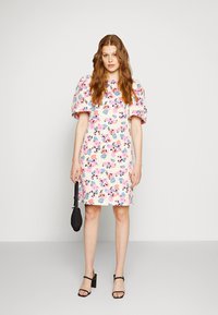 Vestido floral em tecido suave e leve com mangas bufantes; apresenta flores em rosa, azul e preto sobre um fundo bege. Sapatos de salto pretos e uma bolsa.
