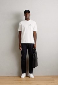 Hvid bomulds t-shirt med en lille blå grafik, mørke denim jeans, hvide sneakers og en sort tote bag. Kortskygget sort cap.
