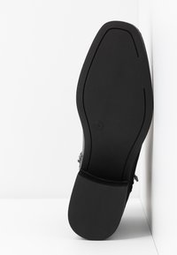 Semelle de chaussure noire avec une surface texturée antidérapante et un petit logo circulaire près de la voûte plantaire, photographiée à la verticale sur un fond blanc d'angle.