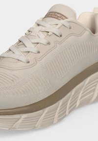 Gros plan sur une basket beige Skechers avec une tige en maille texturée, une semelle épaisse à motifs et des lacets blancs.