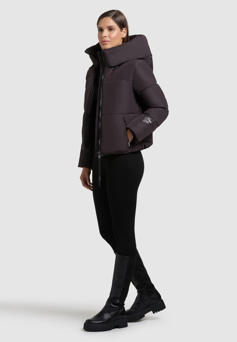 Zwarte pufferjack met een hoge kraag, ritssluiting en elastische manchetten; gedragen met zwarte leggings en platte kniehoge laarzen.