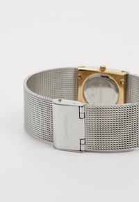 Ezüst hálós karóra téglalap alakú aranytokkal, "Calvin Klein" felirattal gravírozott csattal és átlátszó hátlappal.