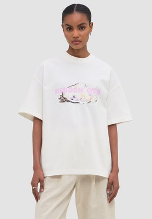 Kobieta o ciemnych włosach w białej, oversize koszulce z ostrygą i różowym napisem „HIDDEN GEM”, nosząca jasnobeżowe spodnie.