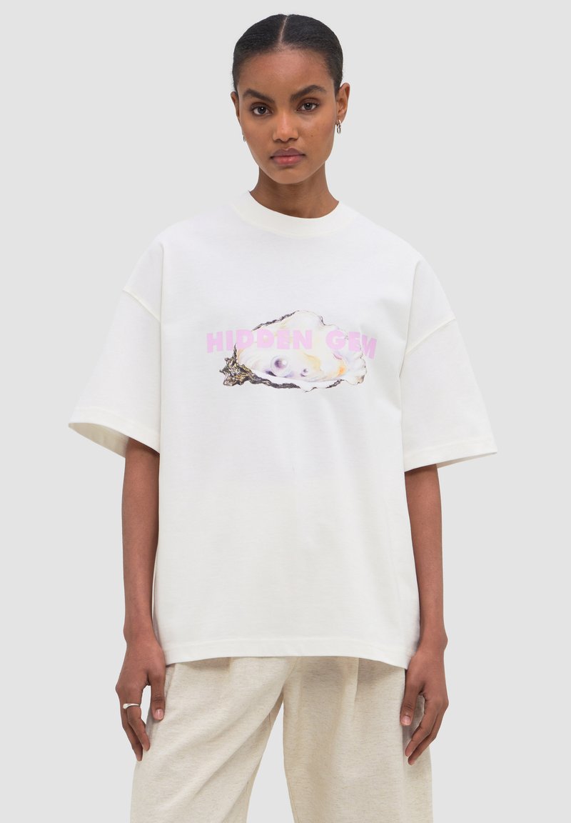 Γυναίκα με σκούρα μαλλιά σε άσπρο φαρδύ T-shirt με στρείδι και το κείμενο «HIDDEN GEM» σε ροζ, φοράει ανοιχτό μπεζ παντελόνι.