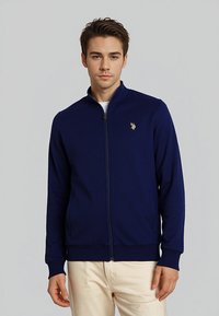 Námornícky zipsový kabátik z hladkej tkaniny s vysokým golierom, dvoma postrannými vreckami a malým zlatým logom na ľavej hrudi. Rukávy sú priliehavé.