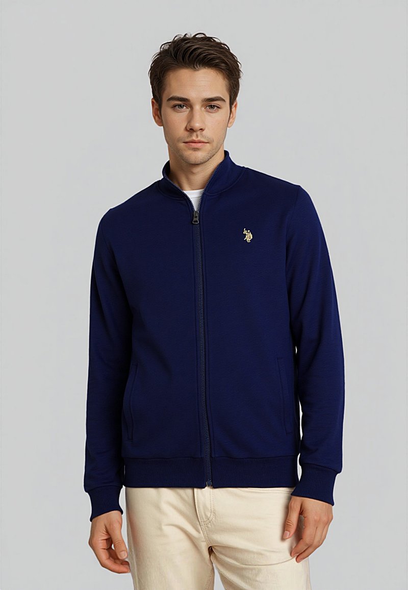 Námornícky zipsový kabátik z hladkej tkaniny s vysokým golierom, dvoma postrannými vreckami a malým zlatým logom na ľavej hrudi. Rukávy sú priliehavé.