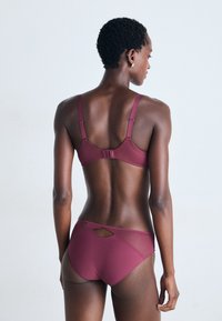Woman with short curly hair wearing a matching maroon bra and panties set, standing with her back to the camera against a plain background.