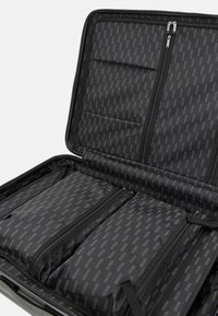 Intérieur d'un sac de voyage noir avec plusieurs compartiments zippés et un motif de logo répétitif. Tissu lisse avec un design structuré.