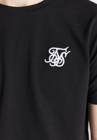 Zwarte katoenen t-shirt met een wit geborduurd logo op de bovenste linkerkant. De stof heeft een zachte textuur en een klassiek ronde halsontwerp.