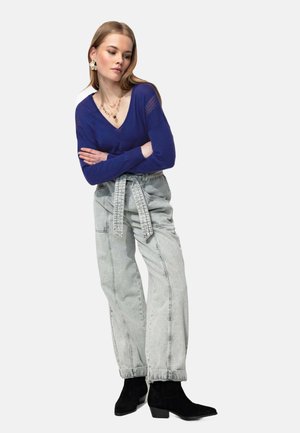 Haut bleu à manches longues en V, jean taille haute gris clair avec ceinture, bottes noires à la cheville élégantes. Les bijoux comprennent des colliers en or superposés et des boucles d'oreilles.