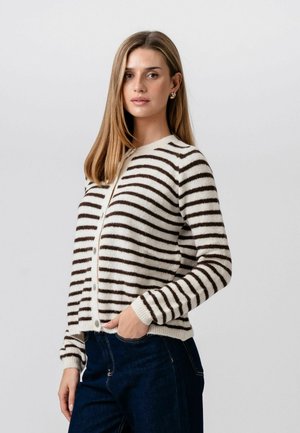 Ung kvinde med glat lysebrunt hår iført guldringe i hoop-øreringe, en hvid og sortstribet knappet cardigan og mørkeblå jeans.