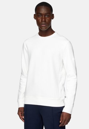 Sudadera blanca de manga larga, con cuello redondo y puños y dobladillo de canalé. Presenta una etiqueta lateral y una textura suave. Combinada con pantalones azul marino.