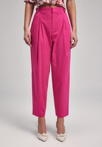 Bright pink high-waisted trousers with pleats, tapered legs, and side pockets. Worn with light-colored, bow-accented heels.